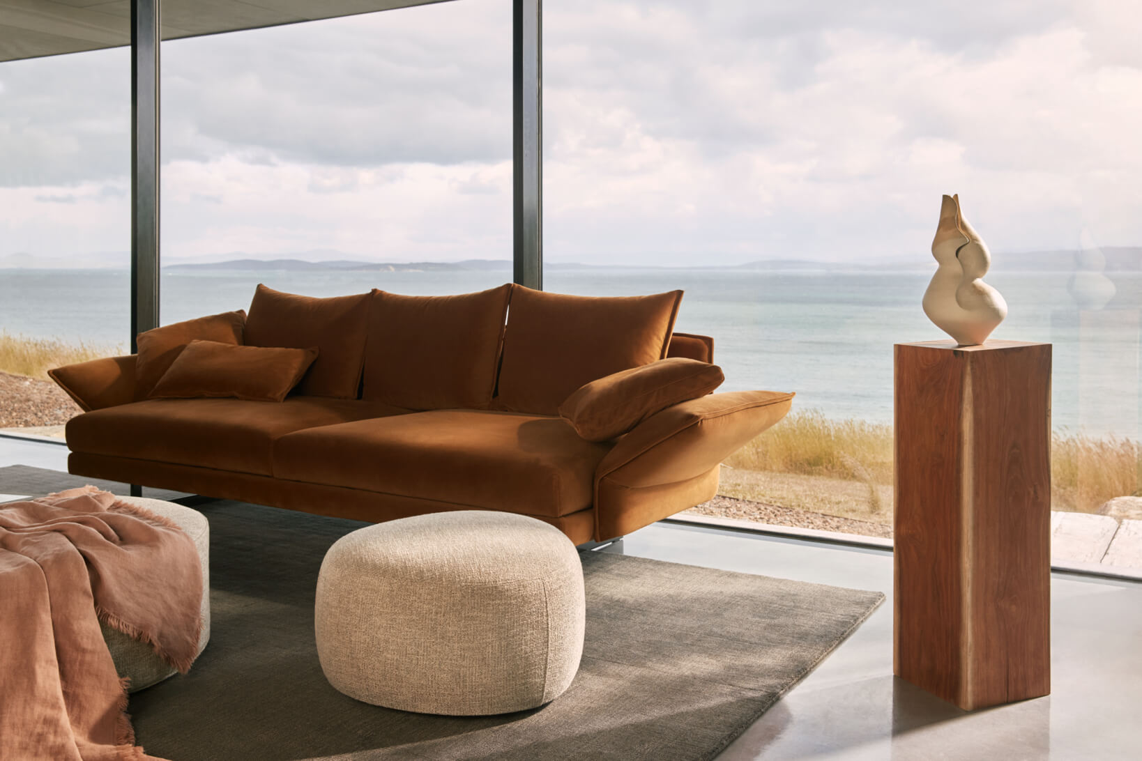 Warm-toned rust velvet sofa with plush cushions beside a beige ottoman in a light-filled contemporary and coastal living room.
